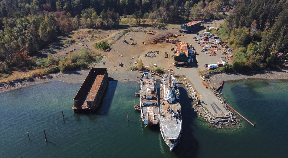 An aerial photo shows an overview of the Deep Water Recovery site. There are two large boats anchored in the water and one barge that is partially on shore and partially in the water. There is a jetty, and a rusty ship appears to be being dismantled on land.