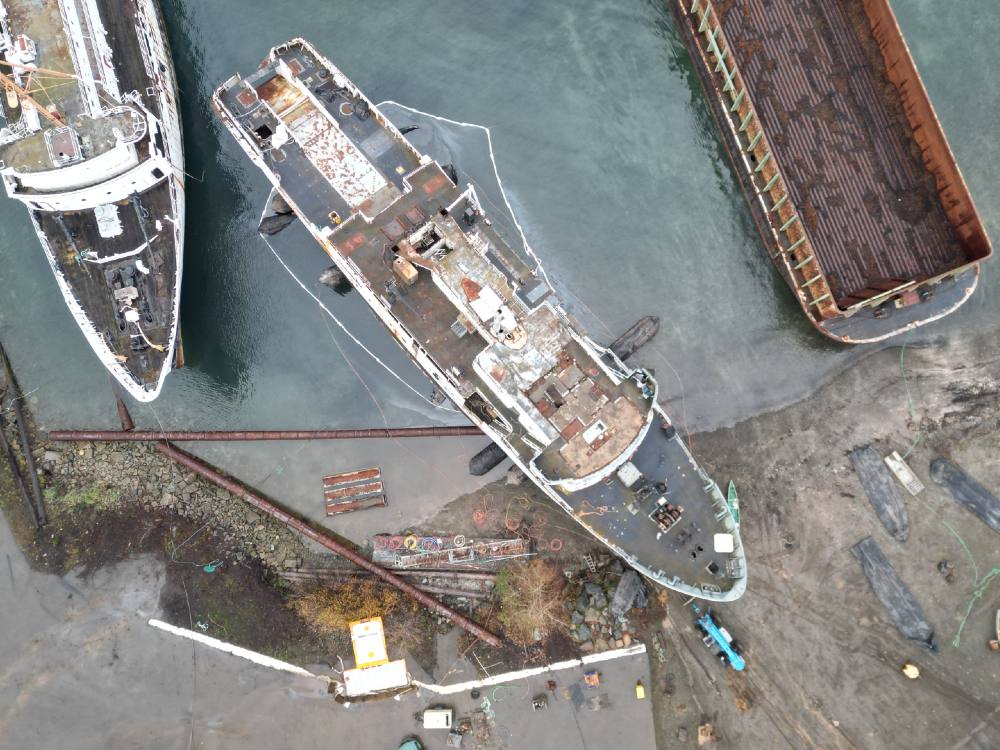 An aerial view looks down at two ships and one barge moored against a pebbly beach. The middle boat has what appears to be an oil sheen surrounding it and a ring of white oil booms. The oil sheen is visible outside of the white oil booms. 
