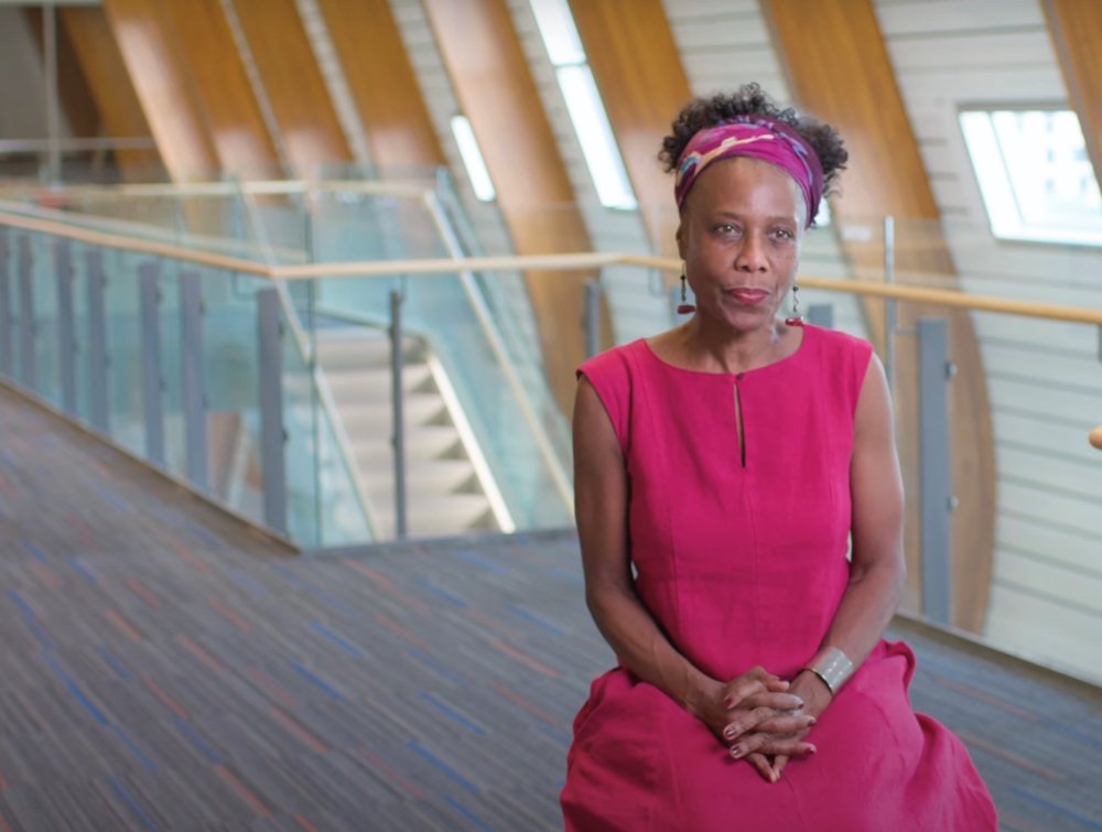 Annette Henry is a Black woman sitting on a chair in an indoor learning space at the University of British Columbia. She is wearing a fuchsia dress and has a colourful scarf in her hair. She is looking to the right of the frame and smiling slightly.