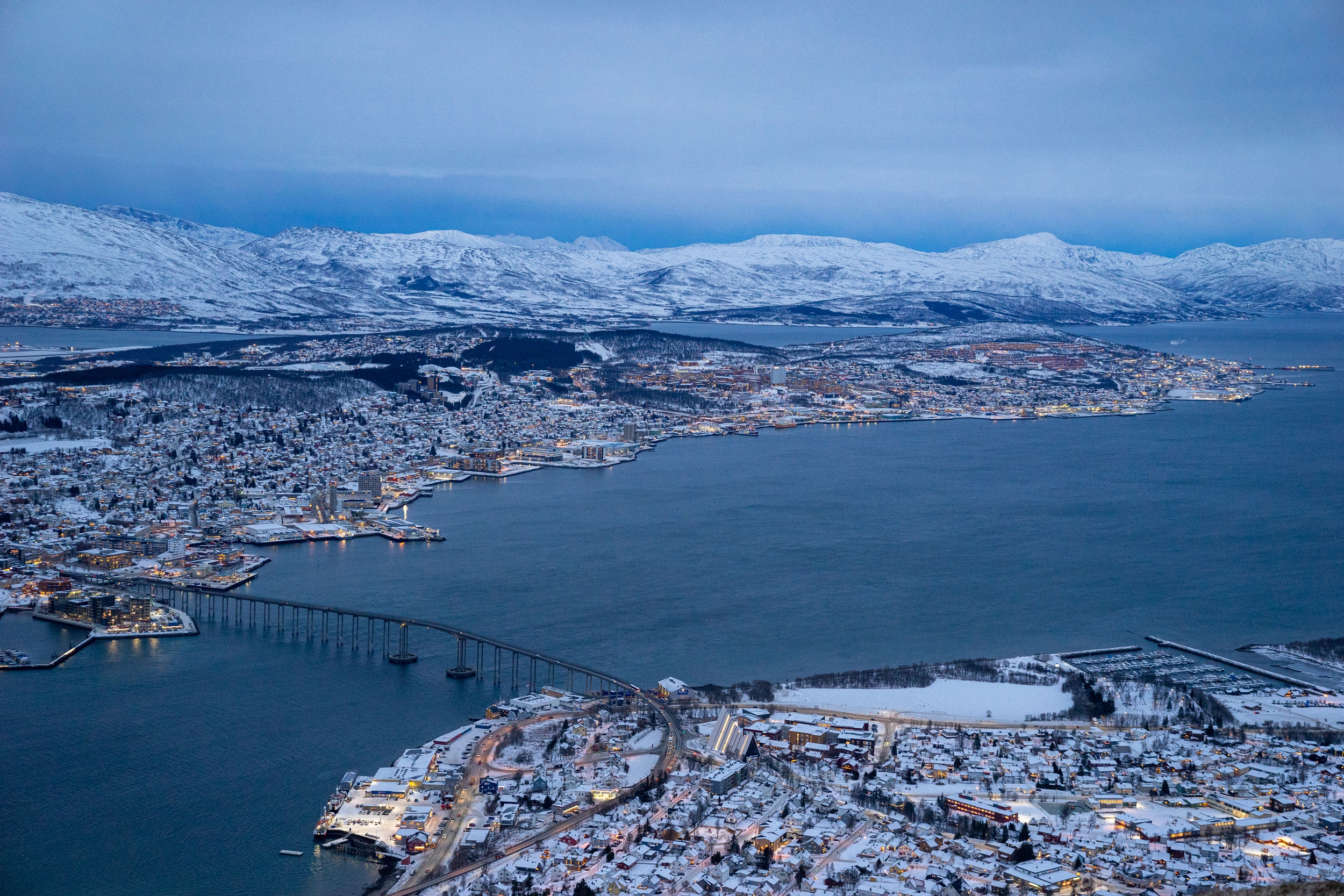 An aerial view of a snowy city on the banks of a large body of water. A bridge connects two parts of the city.
