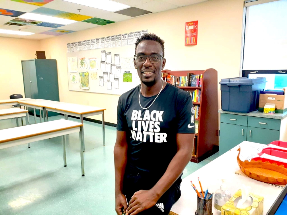 Michael Musherure is a Black man in glasses, looking at the camera with a friendly expression on his face. He is wearing a black T-shirt that says “Black Lives Matter.” He is leaning on a desk in a high school classroom with a green floor.