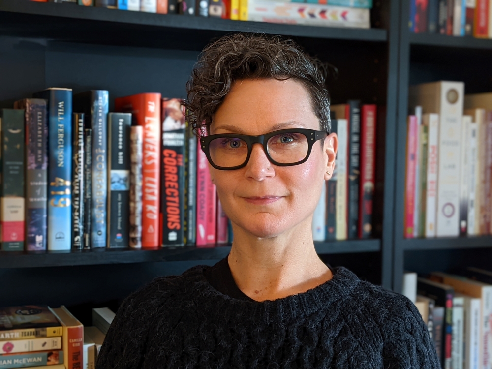 Melanie Scheuer is a white woman with short dark hair. She is standing in front of a bookshelf wearing dark glasses with thick frames. She is looking at the camera and smiling slightly.