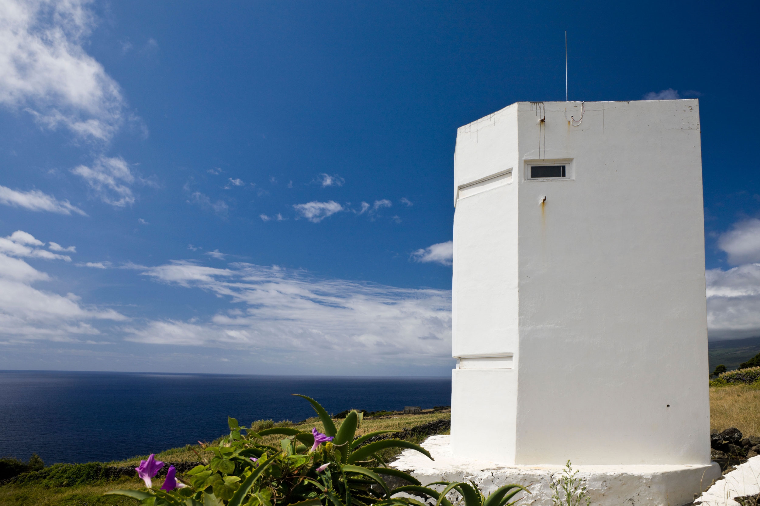 A white tower rises out of the grass into the sky, sort of looking like a cross between a lighthouse and a military building. It is relatively close to shore.