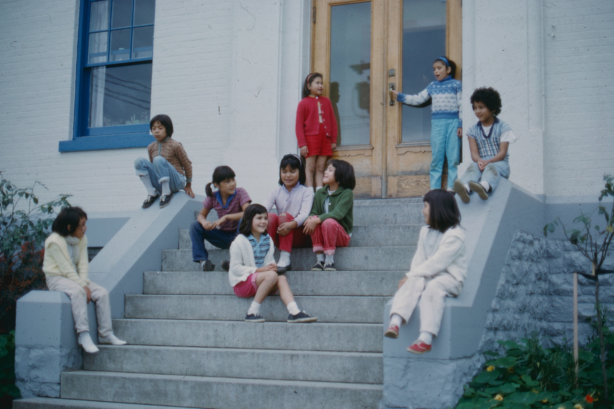 Ten children, all young, are sitting on wide steps that led to a white brick building.