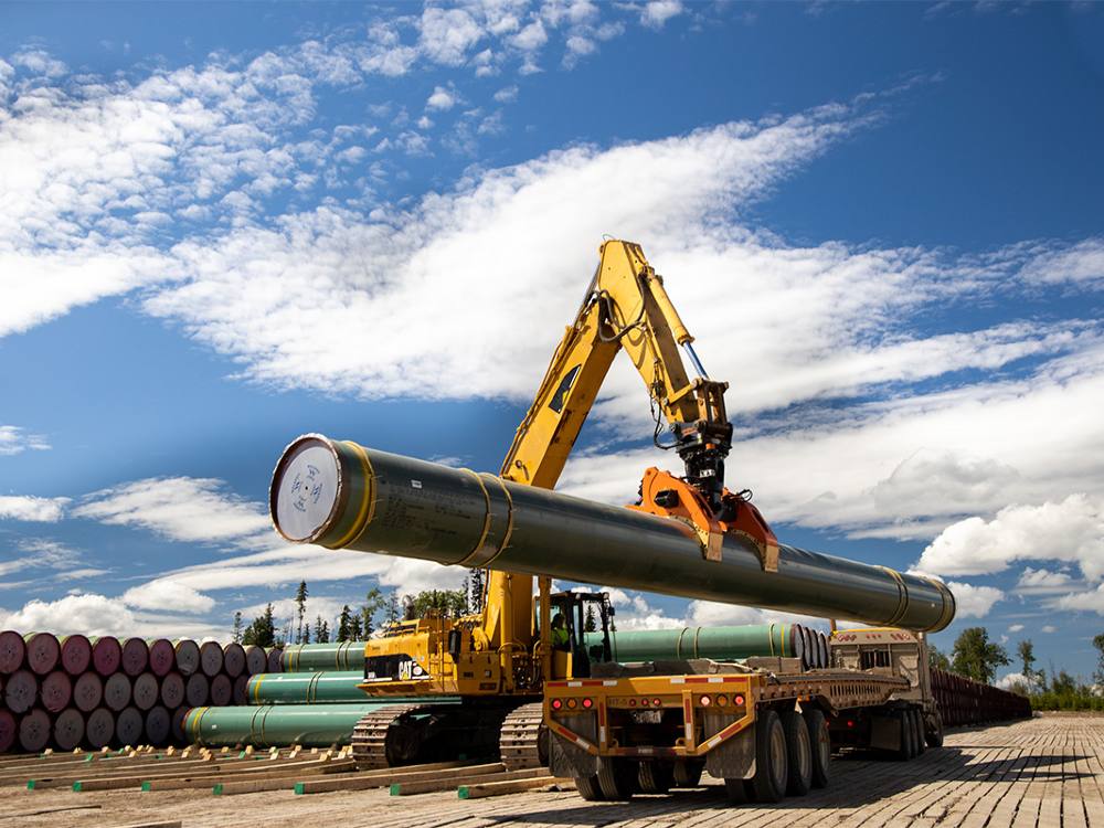 A large yellow machine picks up a section of green metal pipe and places it on a flatbed truck. Sections of green metal pipe are stacked behind.