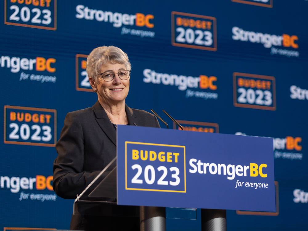 A person wearing a black suit with greying hair smiles at the camera, standing at a podium that says "Budget 2023: Stronger BC for Everyone."