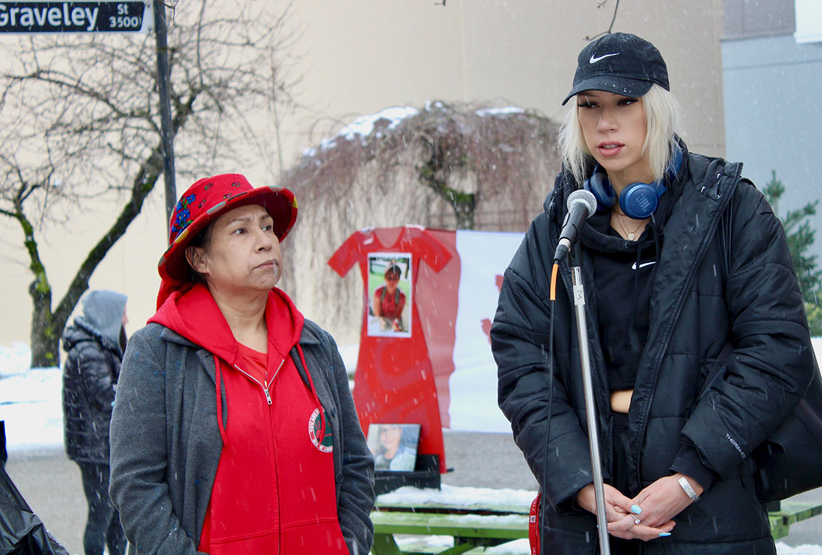 A woman in a red hoodie and hat grey jacket looks toward a taller blonde person wearing black, including a ball cap. In the background, a red dress hangs on a wall.