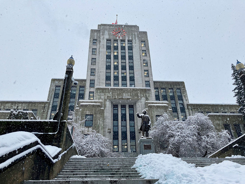 Vancouver City Hall, a large grey stone office building, is shown under a pale blue sky. Snow covers part of the wide steps leading to the main doors.