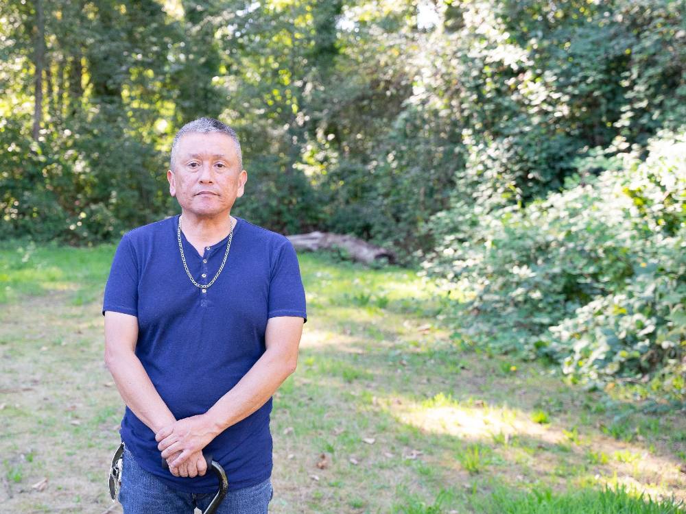 George Chaffee stands to the left of the frame in a blue T-shirt. He has a silver chain around his neck and is holding a pair of sunglasses. He is looking directly towards the camera with a neutral expression. He is standing in a grassy green gove surrounded by deciduous trees on a sunny day.