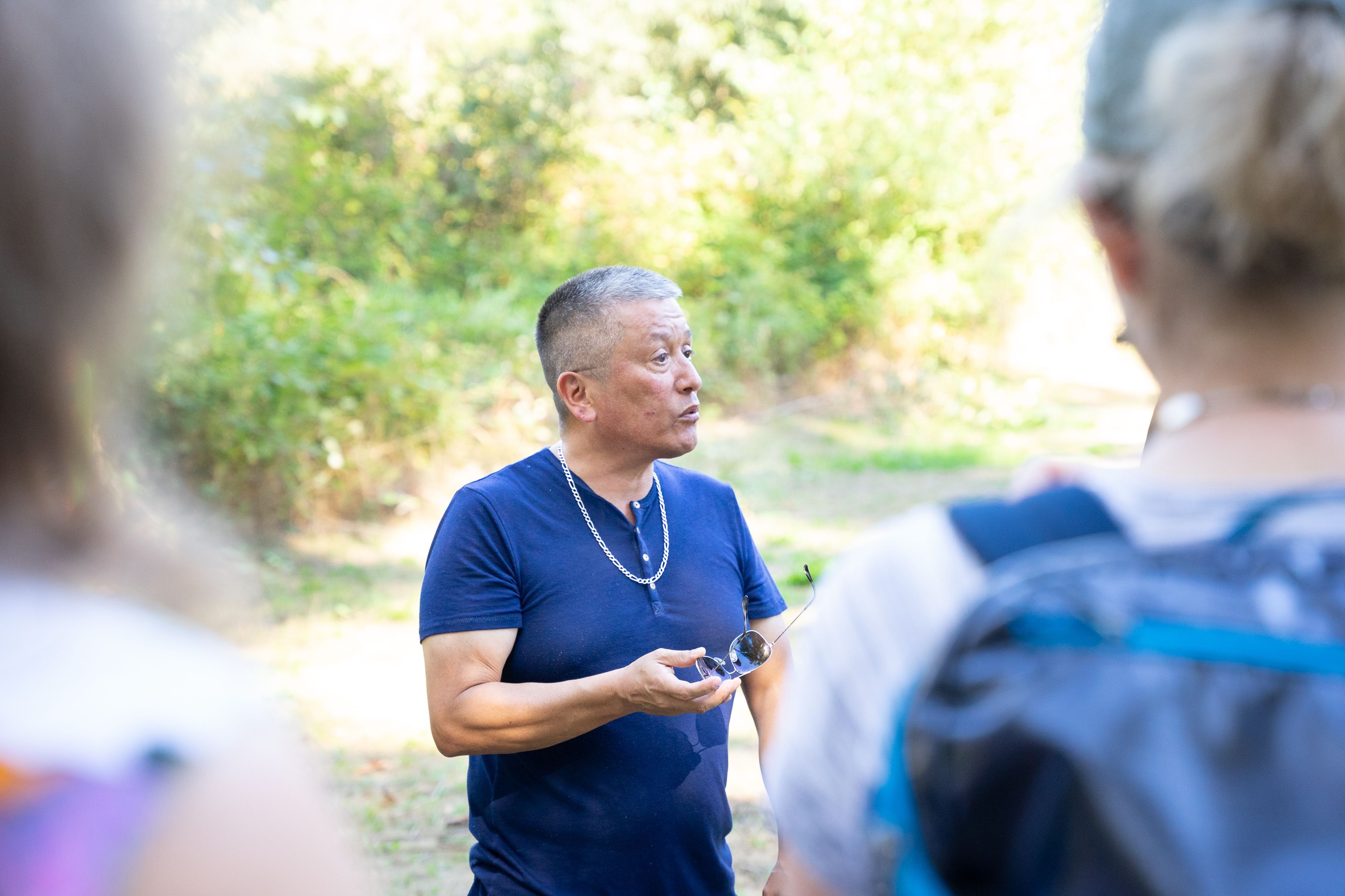 George Chaffee stands looking towards the right of the frame, mid-speech. He is holding a pair of sunglasses in his left hand and is wearing a blue T-shirt. Two people stand in soft focus in the foreground, part of a group he is leading. They are standing in a sunny green grove.