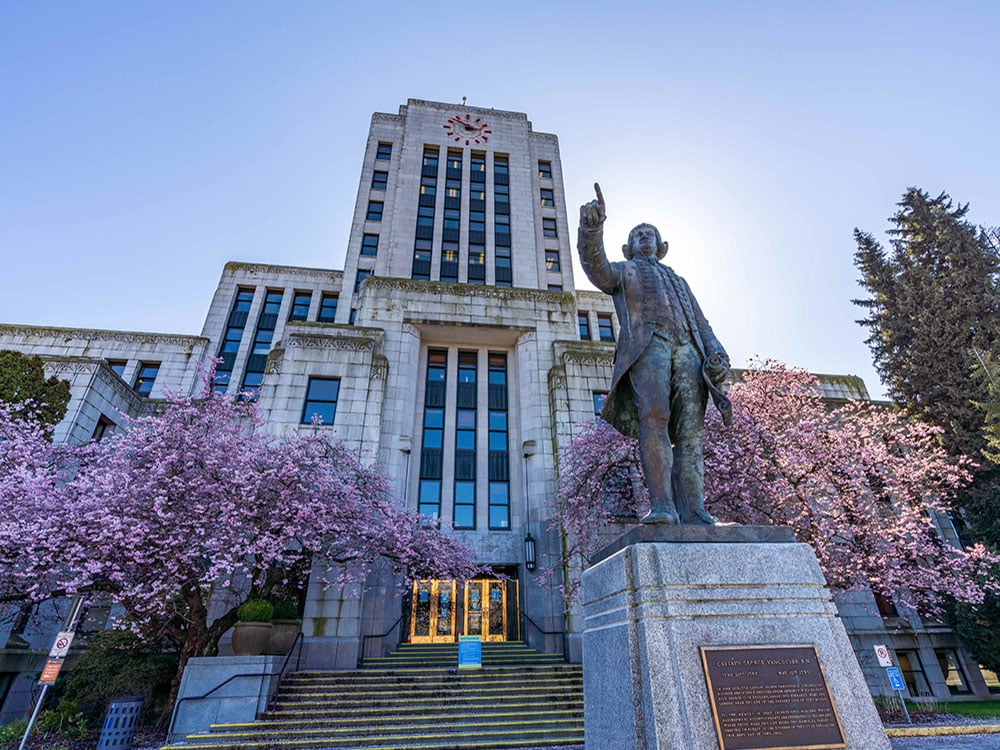 Vancouver City Hall, a large stone building with a clock at its top stands behind blossoming trees. A grey statue of Capt. George Vancouver is in the foreground.