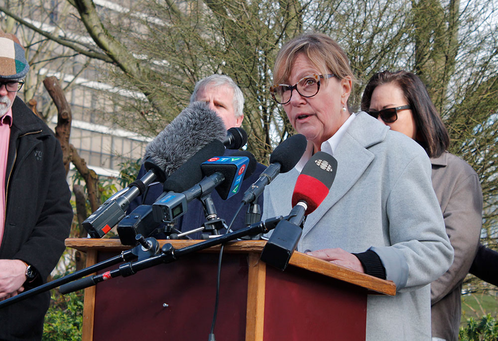 A person with a jacket and wearing glasses stands outdoors at a podium crowded with media microphones.