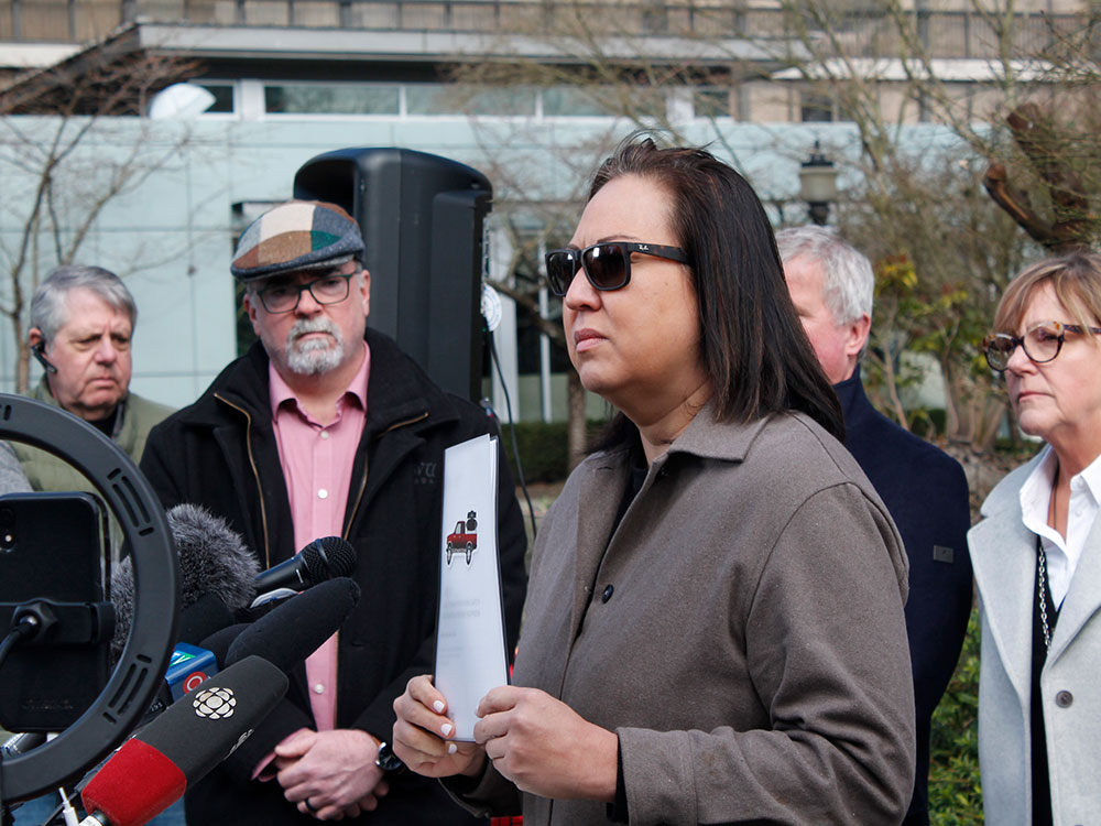 A person with medium-length dark hair, grey jacket and sunglasses stands outside at a podium. She holds a white document as others look on.
