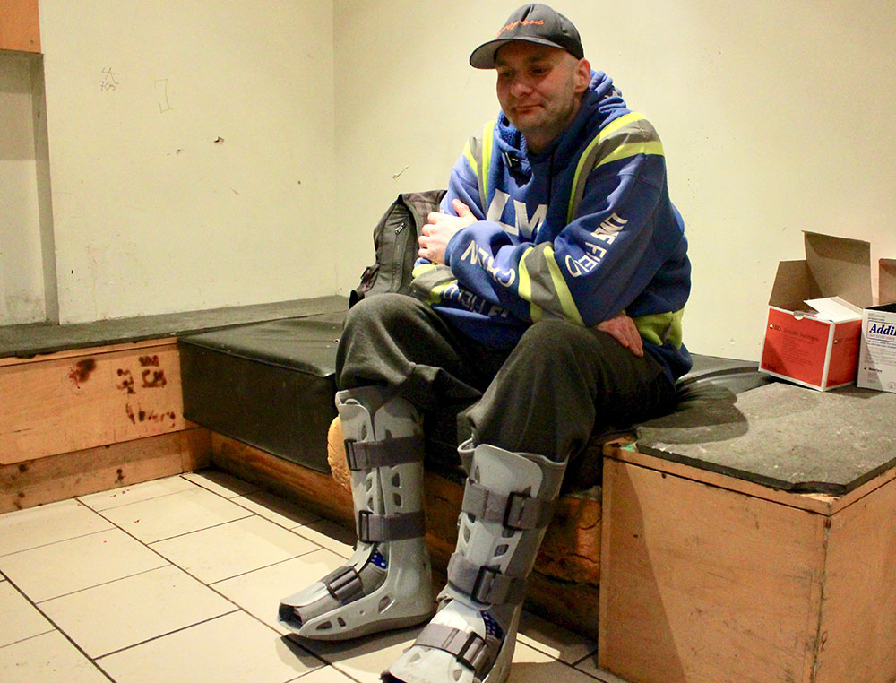 A person sits on a bench in an apartment lobby, wearing a ball cap and has grey plastic braces on his lower legs and feet.