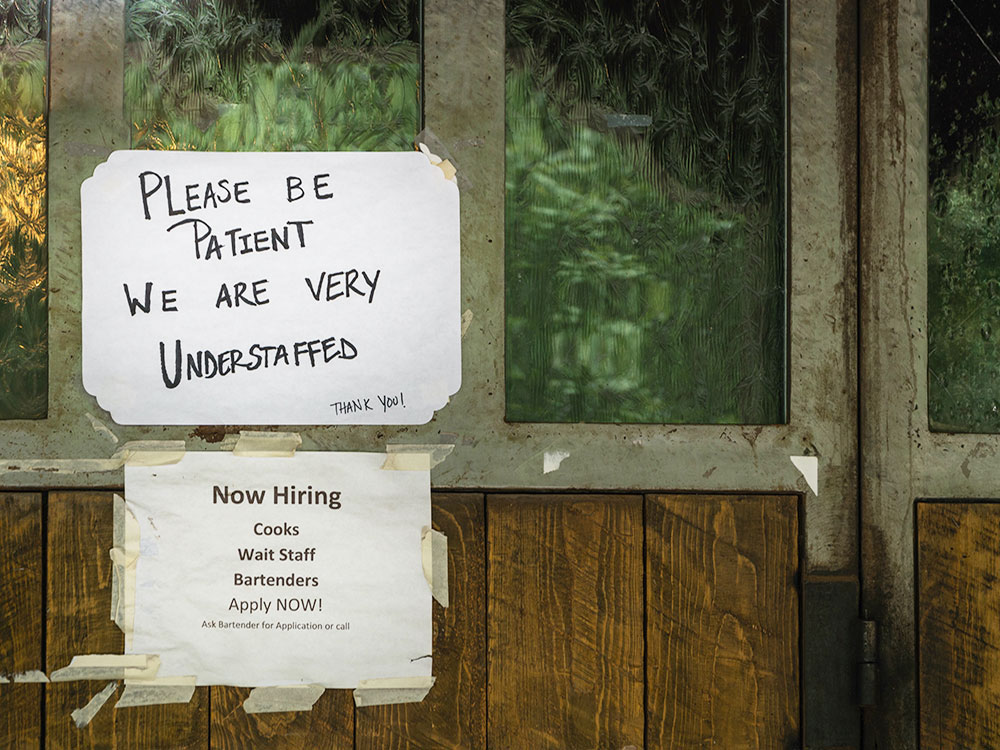 Two hand-lettered signs on a wooden bar door say ‘Please Be Patient — We Are Very Understaffed” and list a list of job vacancies.