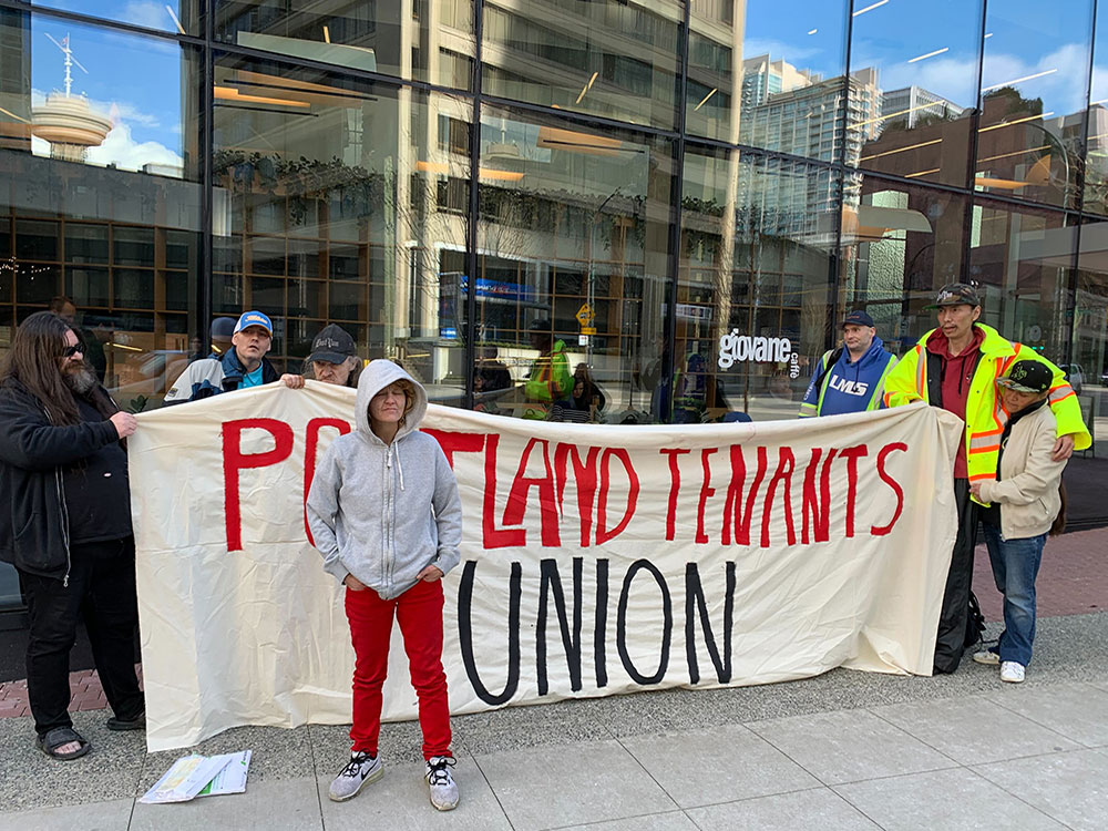 Eight people stand in front of a gleaming office tower holding a large cloth banner that reads “Portland Tenants Union.”