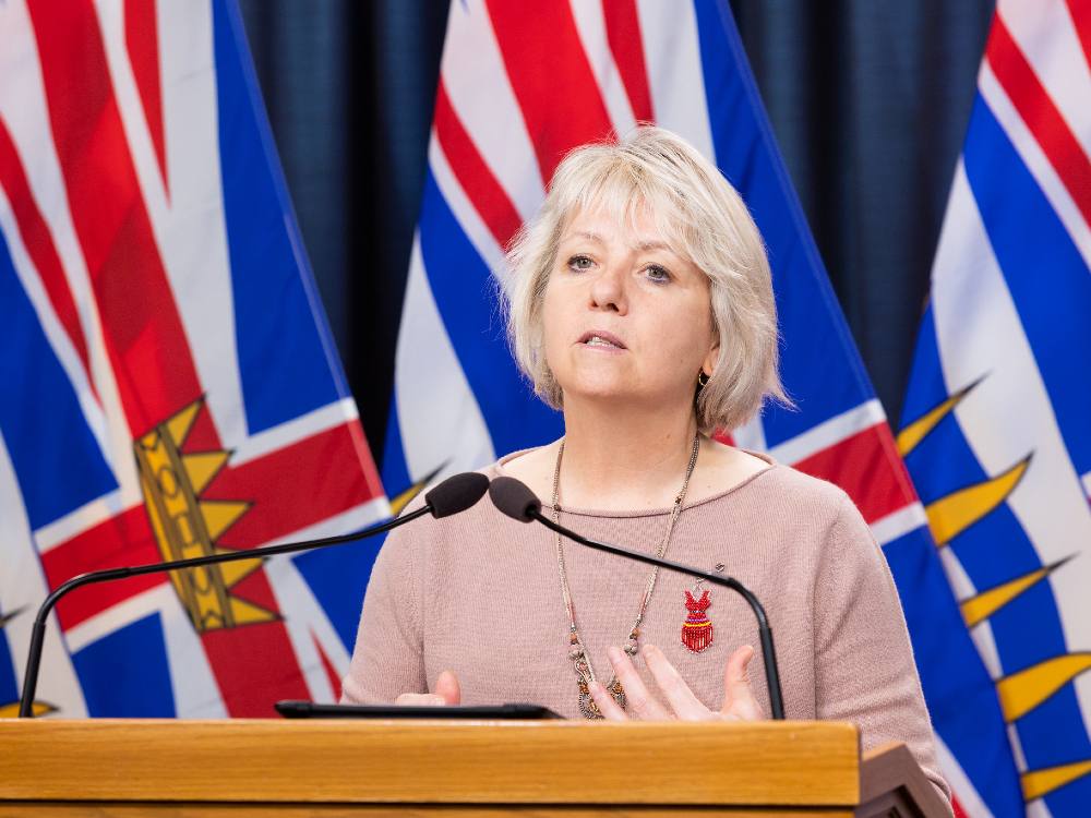 Dr. Bonnie Henry stands in front of a B.C. flag and behind a wooden podium, wearing a pink sweater.