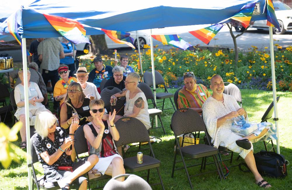 Queer seniors gather outside under a tent on a sunny day to celebrate the annual Aging with Pride event.