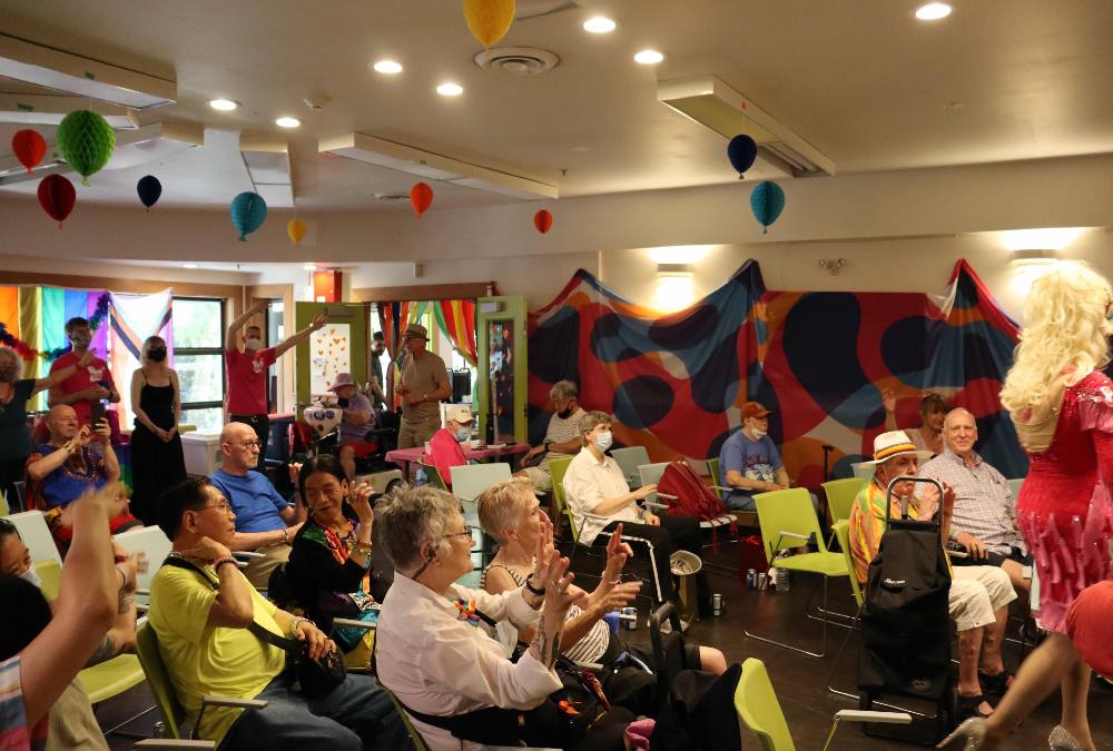 Queer seniors gather in a room decorated with pride flags and other colourful decorations. It looks like there is probably music playing—some people are dancing.
