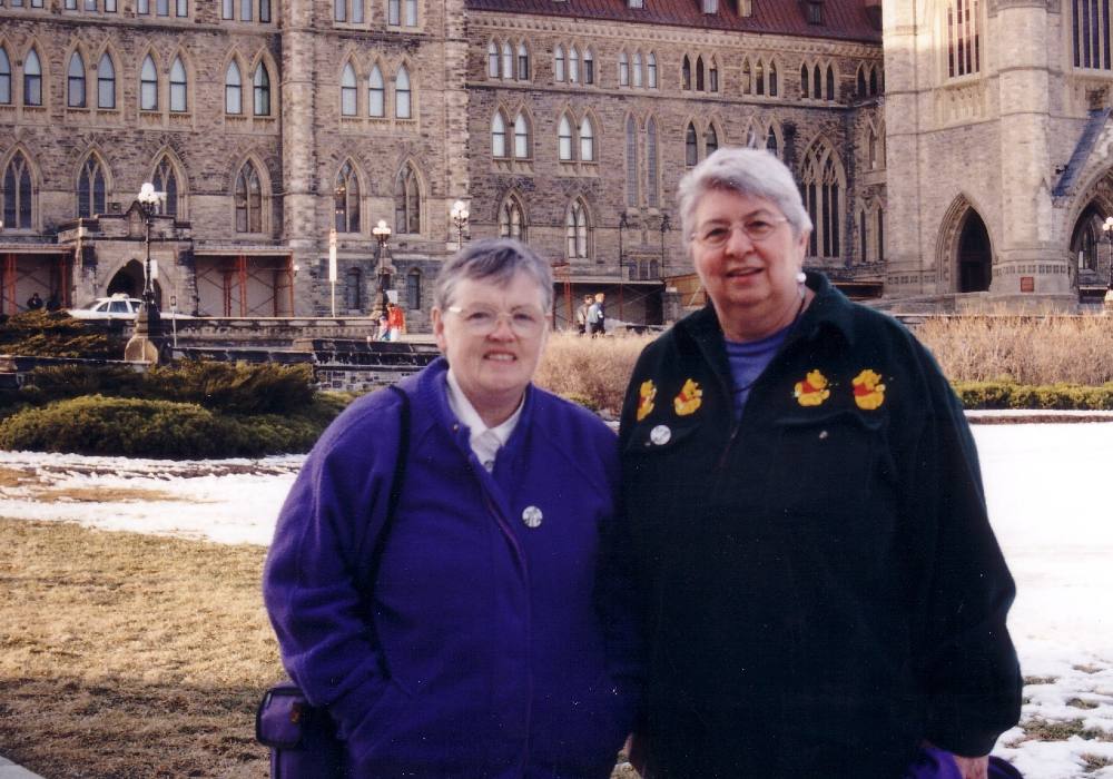 Two women with short grey hair in warm polar fleece coats stand together in front of a large, fancy limestone building in Ottawa.