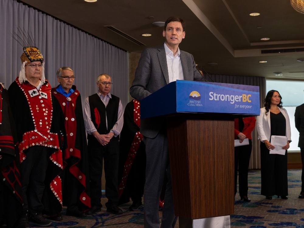 Premier David Eby stands at a podium, with Haisla nation members in traditional regalia behind him. On his left are Haisla Chief Coun. Crystal Smith and Environment Minister George Heyman.