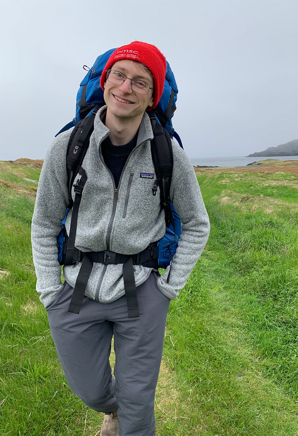 A person wearing a red toque and backpack hikes in a grassy area near an ocean.