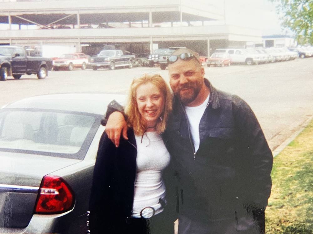 An aged photo shows a young woman with blond hair smiling at the camera. She stands next to a man with a beard and wearing sunglasses on his head, also smiling. They are outdoors and have their arms around each other.
