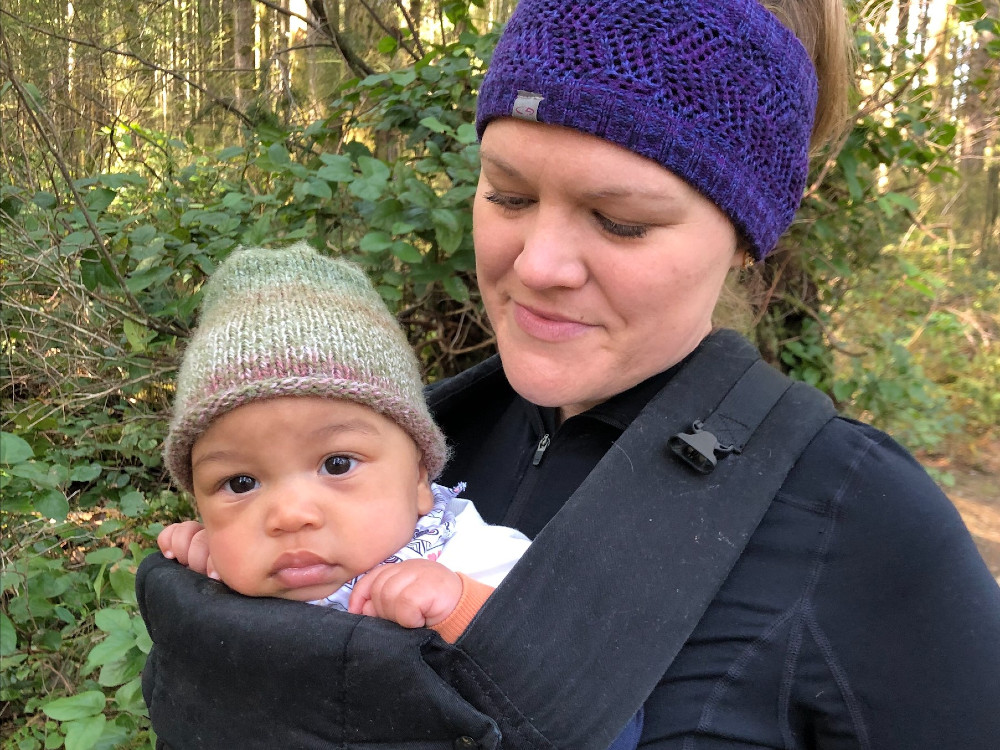 A woman in running gear on a forest path looks down at her child in a chest harness, whose big dark eyes stare at the camera.