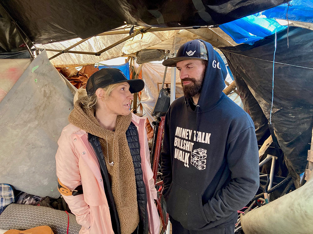 Two people look at each other inside their cluttered shelter, one wearing a ball cap and a pink jacket and large beige sweater, the other a ball cap and blue hoodie.
