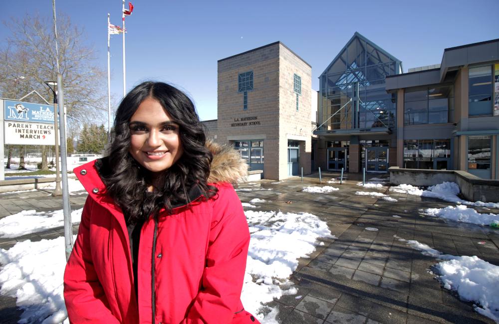A South Asian female teen stands in front of school buildings with snow on the ground. A sign identifies the school as L.A. Matheson Secondary.