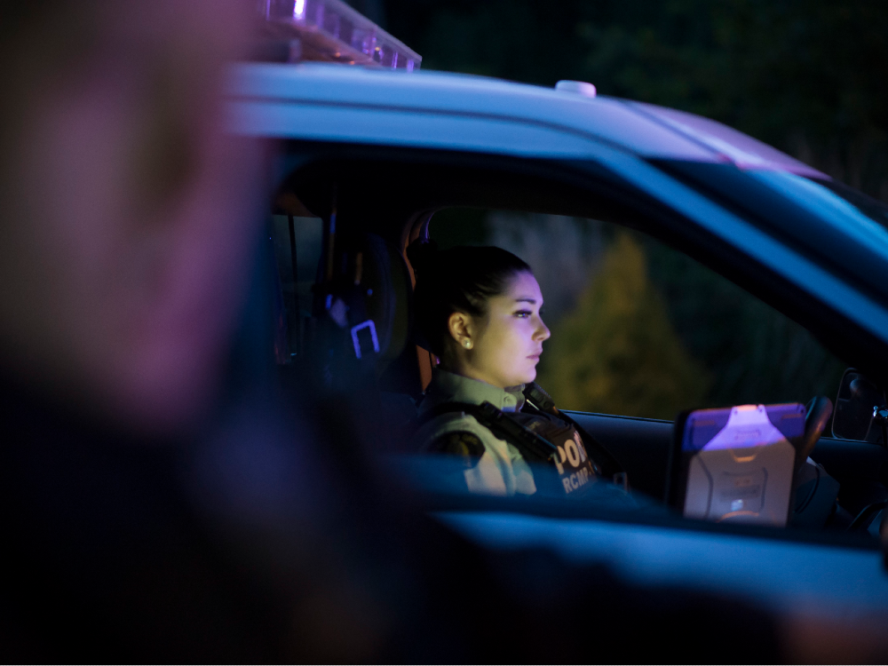 A brown-skinned, dark-haired female police officer sits in a police vehicle at night.