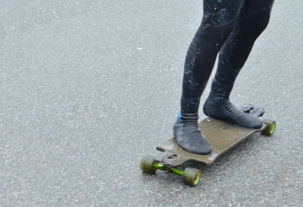 A man wearing a black and blue wetsuit rides a longboard over grey cement. 