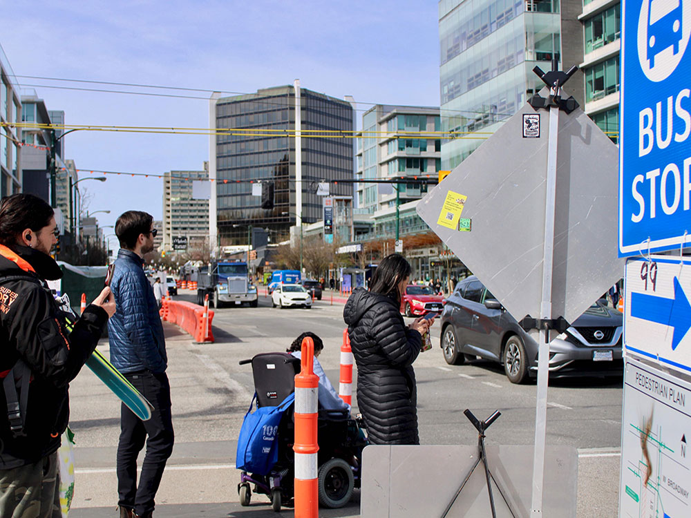 Four people stand waiting to cross the street at a busy Broadway intersection. Pylons and plastic barriers show evidence of construction amid traffic.
