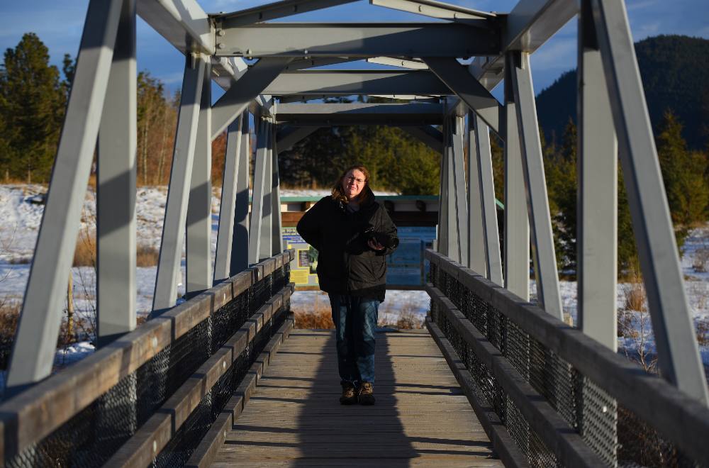 A woman, Billie Sheridan also shown in the main photo at the beginning of the story, looks at the camera in a big coat as she stands on a bridge constructed of metal beams.