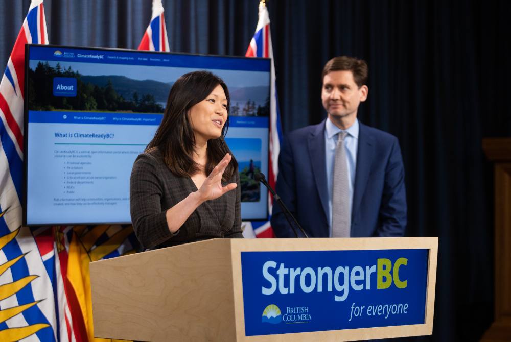 A thirty-something woman of Chinese heritage, Minister Bowinn Ma, speaks at a podium with a tall white man, Premier David Eby, looking on.