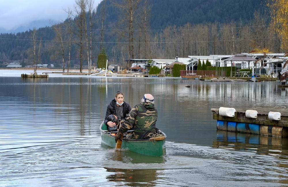 In the top photo smoke pours over a ridge behind a building with parked cars in front. In the bottom photo a young woman with long brown hair and a worried expression on her face is paddled in a canoe by a middle aged man with his back to the camera.