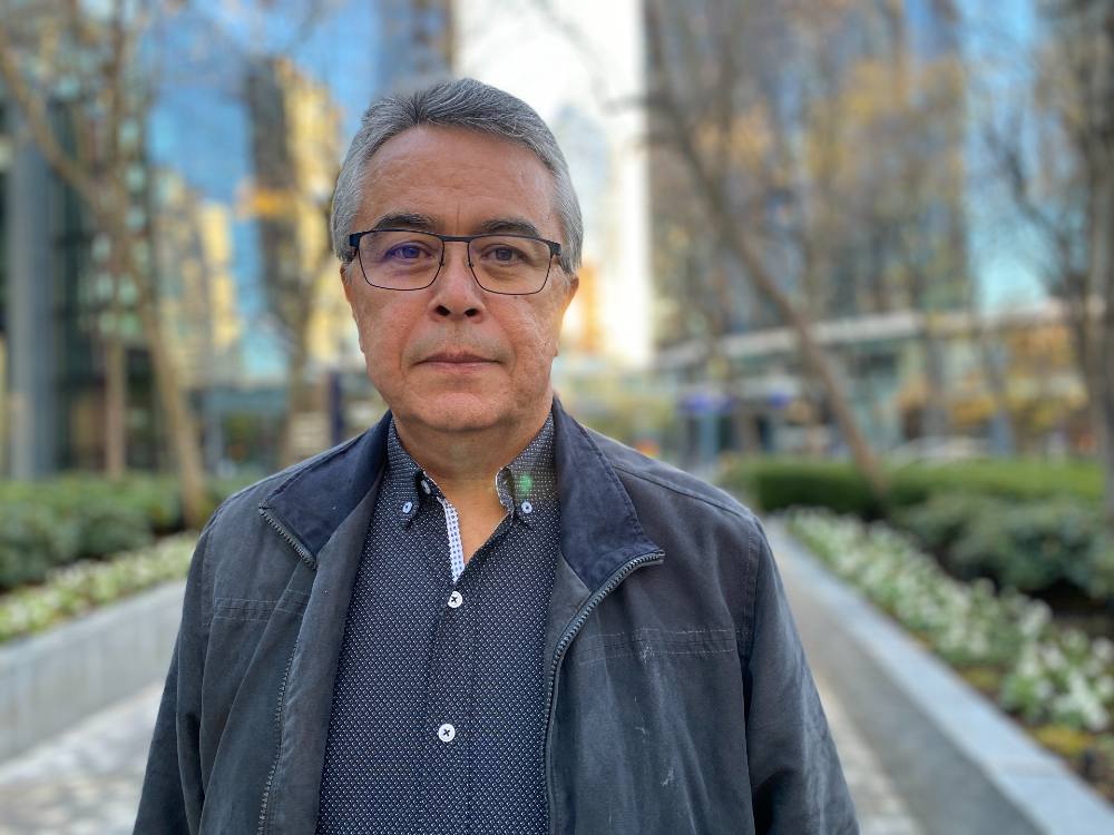 A middle aged man in a black jacket stands outside in an urban setting. He is Indigenous.