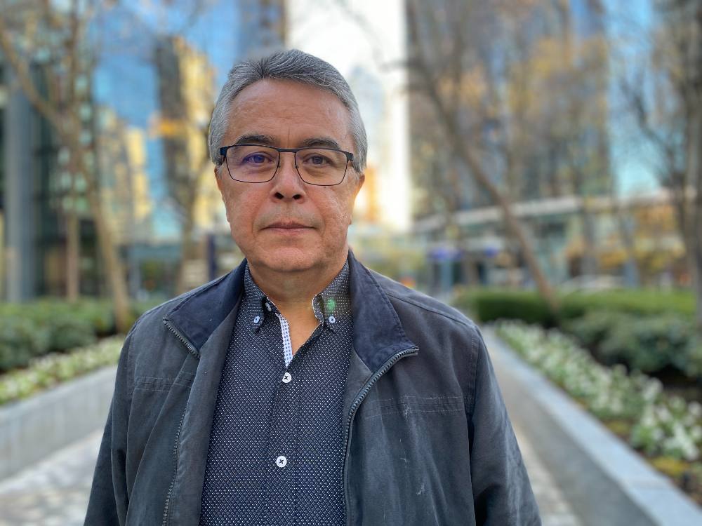 A man stands on a cement pathway in front of an urban setting of glass buildings. He is wearing glasses, a button-up navy shirt and blue jacket.