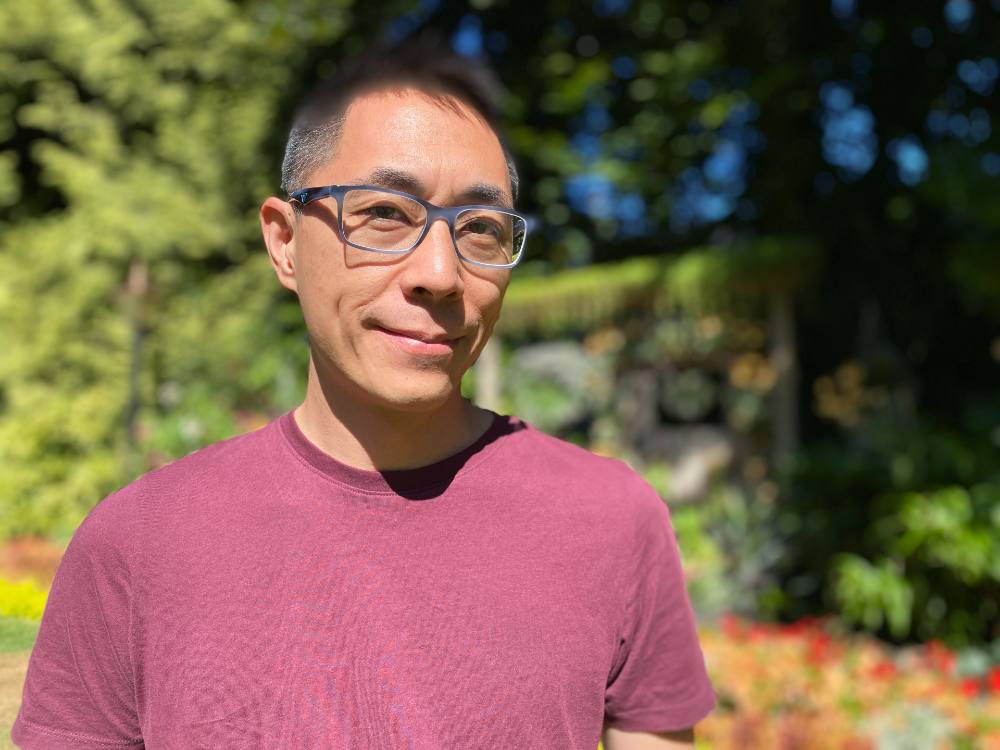 A man of Chinese heritage with black-rimmed glasses and a bright red shirt looks at the camera with a small smile.