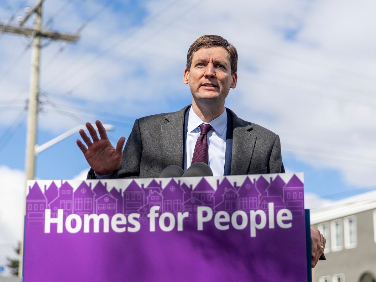 Premier David Eby, in a suit, stands behind a podium that says ‘homes for people.’
