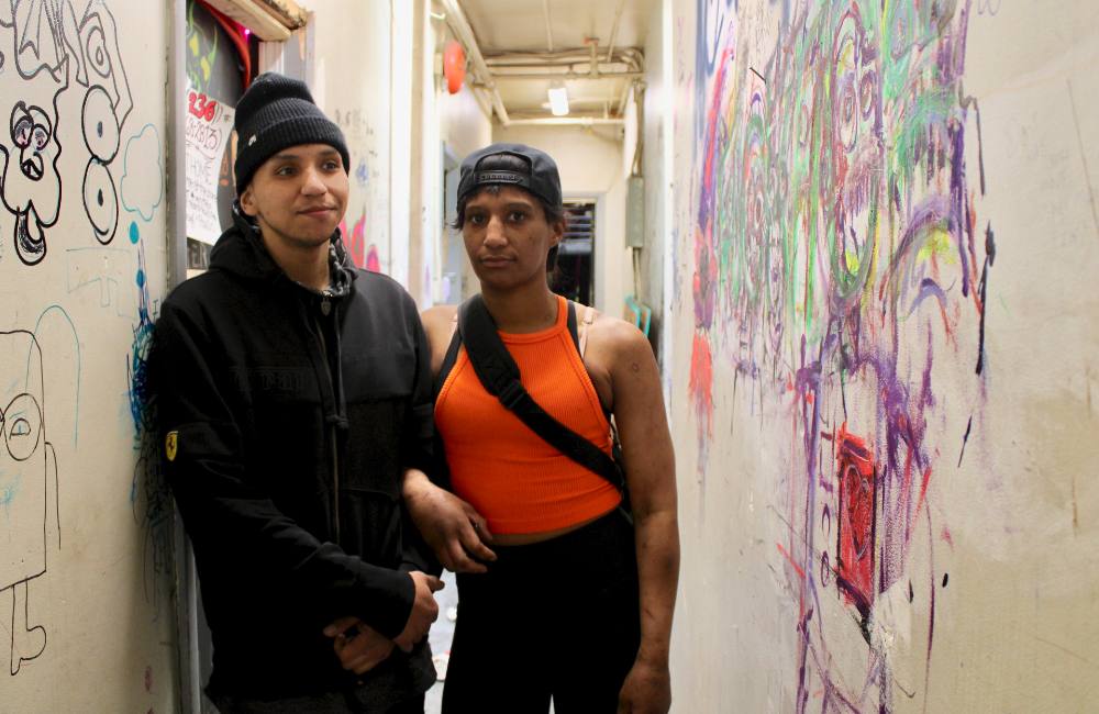 Two people stand a narrow hallway, covered with graffiti.