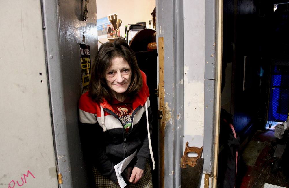 A person stands in the doorway of an SRO room, wearing a nylon jacket, a little hunched over and smiling at the camera.
