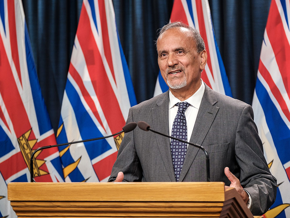 Labour Minister Harry Bains, in a grey suit, white shirt and blue-patterned tie, stands at a podium with flags behind him.