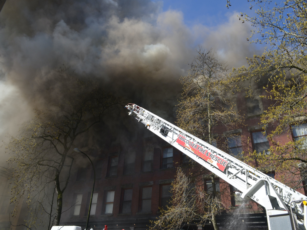A white fire department ladder reaches toward a brick building, as large amounts of dark smoke pour from the upper floors.