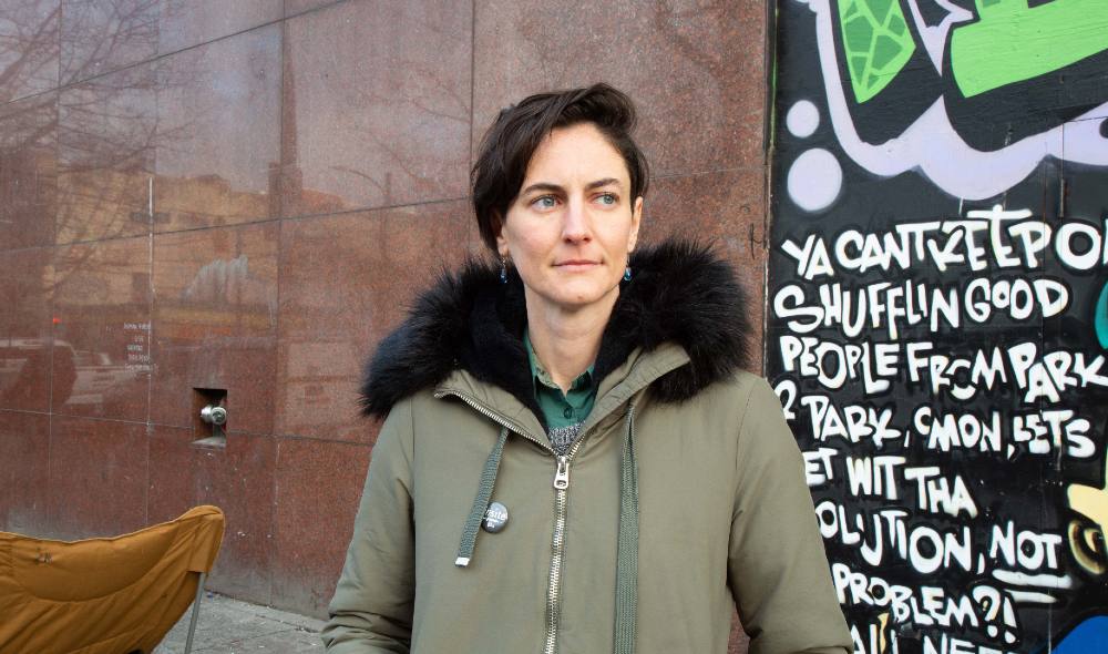 A person with short, swoopy hair, wearing a parka, earrings and a pin saying “insite,” stands in front of a mural.