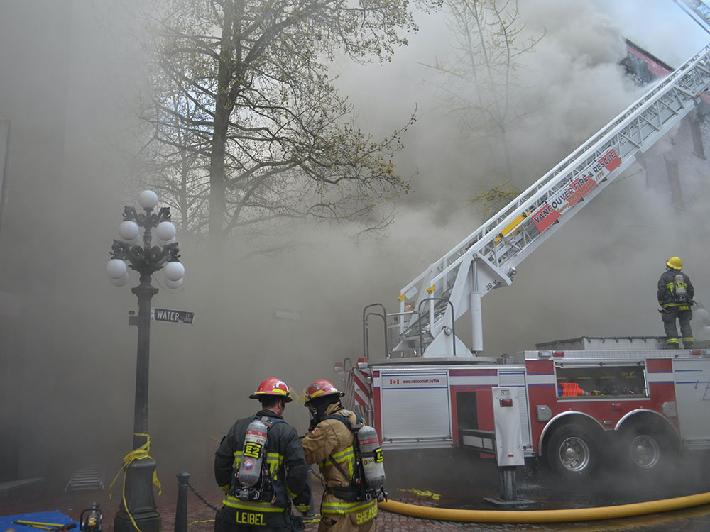 Firefighters wearing full protective gear and oxygen tanks stand near a fire truck with its white ladder raised as clouds of smoke swirl around them.