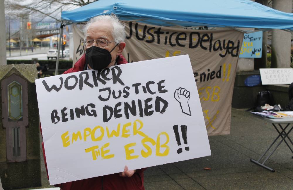 A person wearing a mask is holding a sign that says ‘Worker Justice Being Denied. Empower the ESB.'