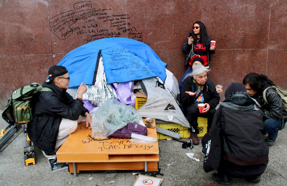 Six people are gathered around a blue tent on East Hastings Street.