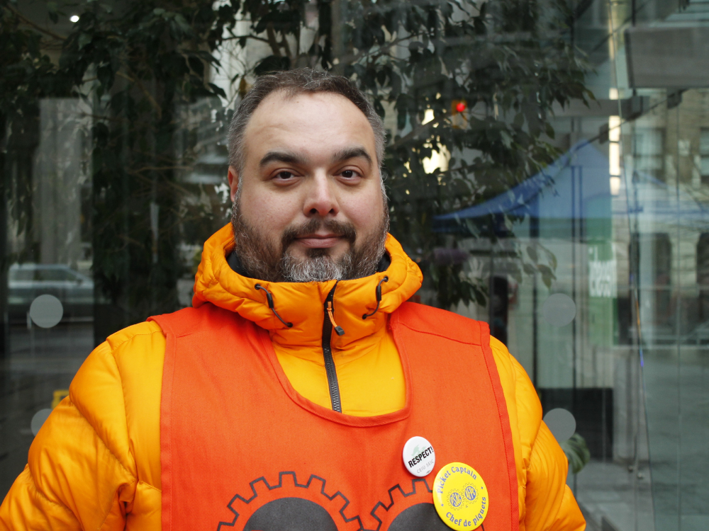 A person with a beard, wearing a yellow parka and orange safety vest smiles at the camera, wearing buttons that say “RESPECT” and “Picket captain.”
