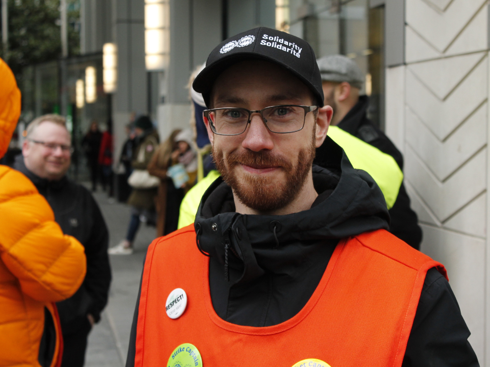 A person with a beard, wearing a black jacket, orange safety vest and black hat saying 'Solidarity.'